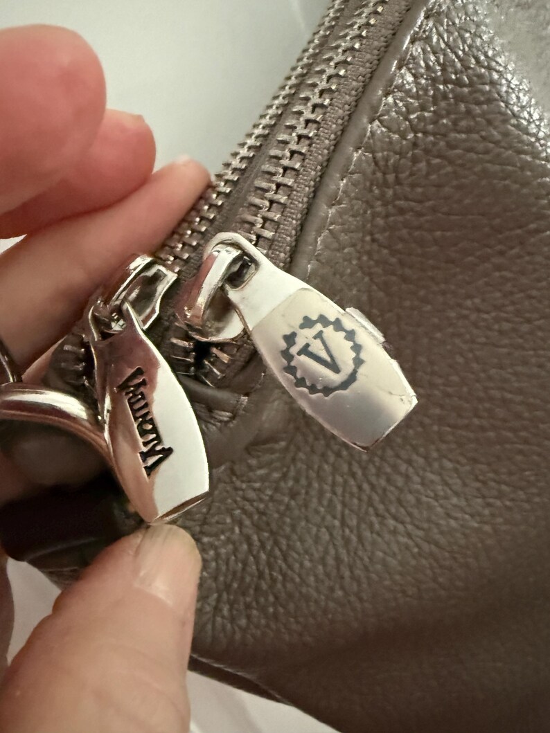 May include: Close-up of a grey leather handbag with a silver zip and zip pulls. The zip pulls have the brand name "Vagrant" engraved on them. The handbag has a textured surface.