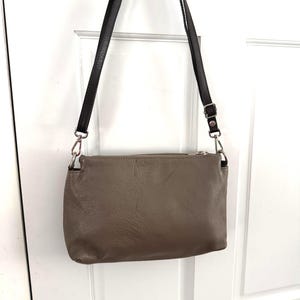 May include: A taupe leather crossbody bag with a black strap. The bag is rectangular with a zip closure and silver-toned hardware. The strap is adjustable and attached with metal rings. The bag is designed to be worn across the body.