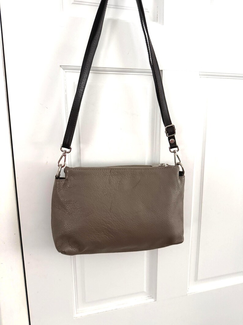 May include: A taupe leather crossbody bag with a black strap. The bag is rectangular with a zip closure and silver-toned hardware. The strap is adjustable and attached with metal rings. The bag is designed to be worn across the body.