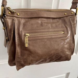 May include: A brown leather crossbody bag with a long adjustable strap. The bag features a front flap pocket and a zippered main compartment. Gold-toned hardware accents the bag, including zippers and studs. The bag is hanging against a white door.