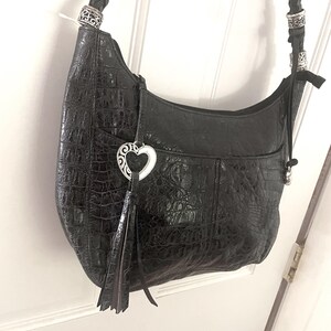 Puede incluir: Bolso de hombro de cuero negro con textura de cocodrilo. El bolso presenta una correa trenzada, herrajes plateados y un charm en forma de coraz&oacute;n con una borla. El bolso est&aacute; colgado de una puerta blanca.