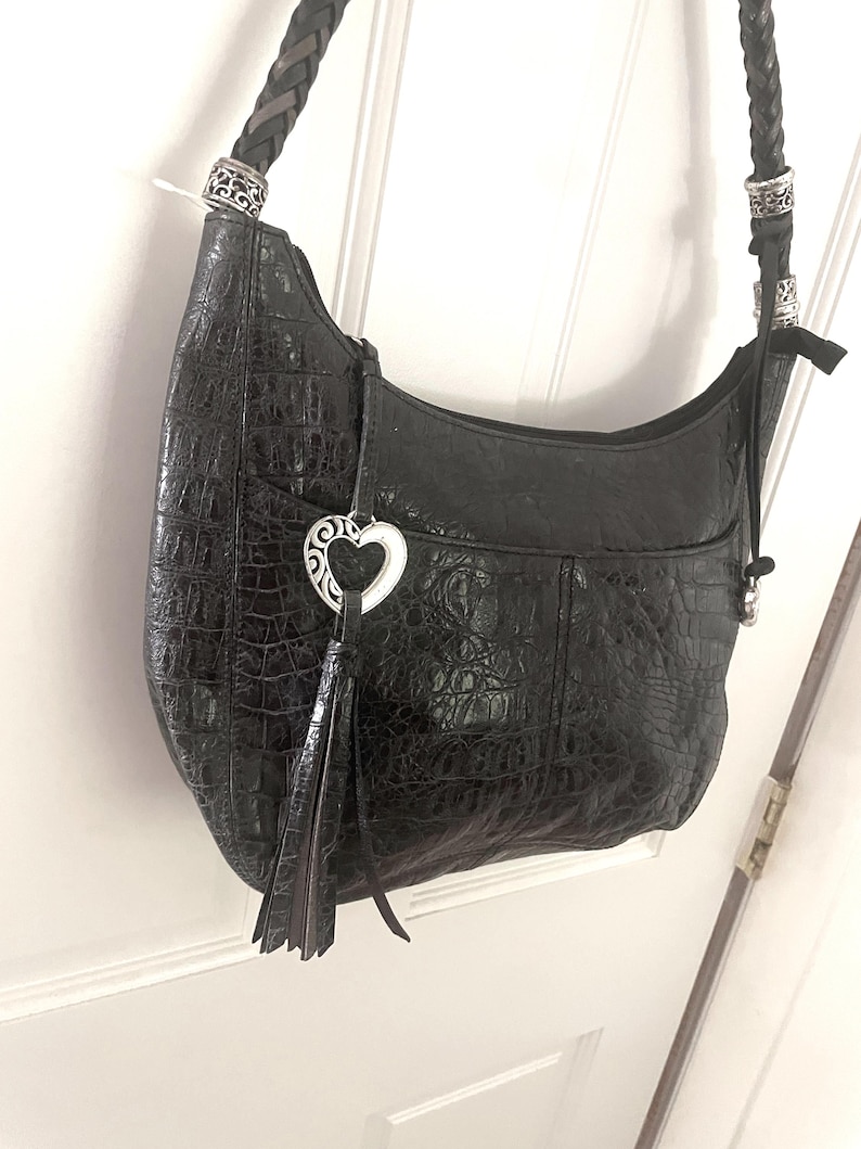 Puede incluir: Bolso de hombro de cuero negro con textura de cocodrilo. El bolso presenta una correa trenzada, herrajes plateados y un charm en forma de coraz&oacute;n con una borla. El bolso est&aacute; colgado de una puerta blanca.