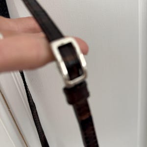 May include: Close-up of a brown leather strap with a silver-colored buckle. The strap appears to be part of a bag or purse, with a textured pattern visible on the leather. The buckle is rectangular.