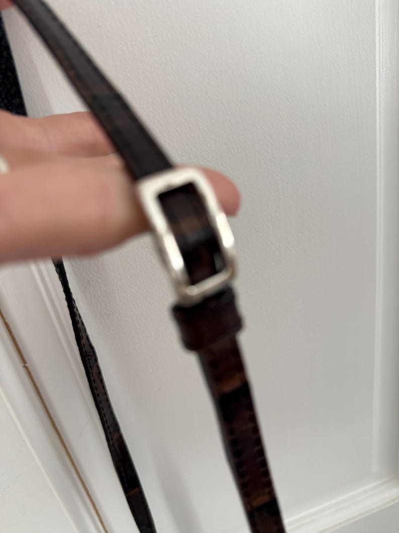 May include: Close-up of a brown leather strap with a silver-colored buckle. The strap appears to be part of a bag or purse, with a textured pattern visible on the leather. The buckle is rectangular.
