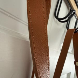 May include: Close-up of a brown leather strap with visible stitching. The strap appears to be part of a bag or accessory, with a textured surface and a warm, natural color. The strap is hanging from a black metal rack.