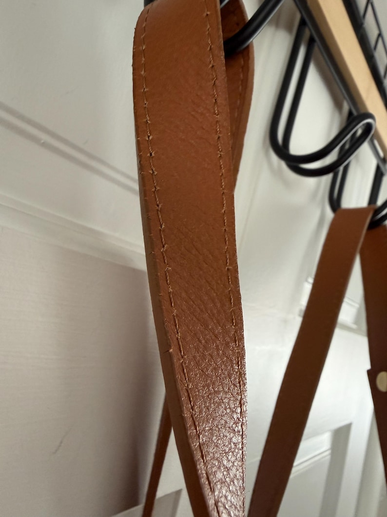May include: Close-up of a brown leather strap with visible stitching. The strap appears to be part of a bag or accessory, with a textured surface and a warm, natural color. The strap is hanging from a black metal rack.