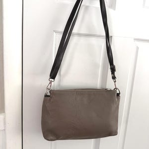 May include: A taupe crossbody bag with a black strap hangs against a white door. The bag has a zippered top and silver-toned hardware. The strap is adjustable and appears to be made of leather.