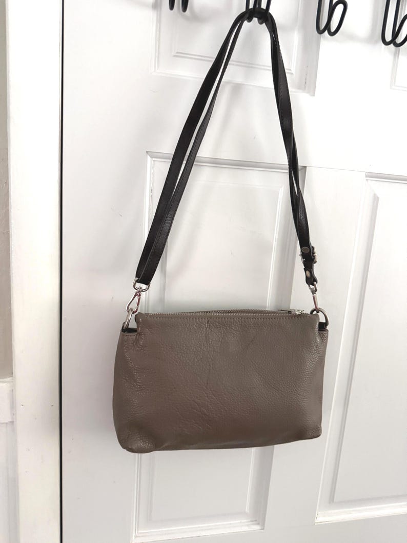 May include: A taupe crossbody bag with a black strap hangs against a white door. The bag has a zippered top and silver-toned hardware. The strap is adjustable and appears to be made of leather.