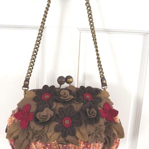 K&ouml;nnte beinhalten: Eine braune und rote Handtasche mit Blumenmuster und Kettenriemen. Die Tasche hat eine tweedartige Stoffbasis und ist mit Filzblumen in Braun- und Rott&ouml;nen verziert. Der Kettenriemen ist goldfarben.