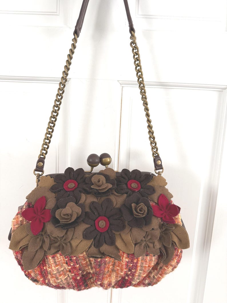 K&ouml;nnte beinhalten: Eine braune und rote Handtasche mit Blumenmuster und Kettenriemen. Die Tasche hat eine tweedartige Stoffbasis und ist mit Filzblumen in Braun- und Rott&ouml;nen verziert. Der Kettenriemen ist goldfarben.