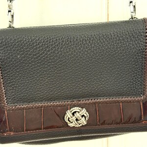 May include: Black leather crossbody bag with a textured finish. The bag features dark brown crocodile-embossed leather accents and a silver-tone knot clasp. A silver chain strap is attached to the top of the bag.
