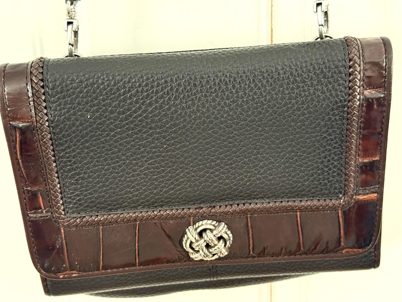 May include: Black leather crossbody bag with a textured finish. The bag features dark brown crocodile-embossed leather accents and a silver-tone knot clasp. A silver chain strap is attached to the top of the bag.