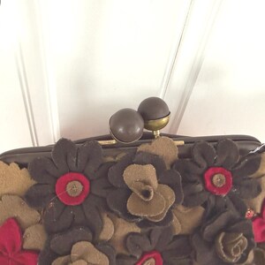 K&ouml;nnte beinhalten: Eine braune Handtasche mit floralem Design. Die Tasche hat dunkelbraune, beige und rote Filzblumen. Sie hat einen goldfarbenen Kettenriemen und einen Verschluss mit zwei braunen Kugeln.