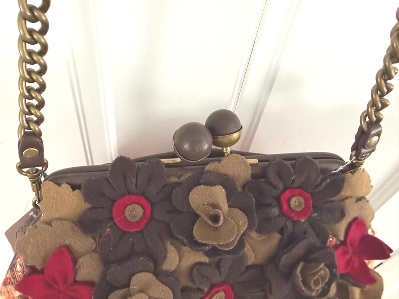 K&ouml;nnte beinhalten: Eine braune Handtasche mit floralem Design. Die Tasche hat dunkelbraune, beige und rote Filzblumen. Sie hat einen goldfarbenen Kettenriemen und einen Verschluss mit zwei braunen Kugeln.