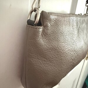 May include: A metallic taupe crossbody bag with a textured leather exterior. It has a silver-tone metal chain strap and hardware. The zipper is visible along the top edge. The bag is hanging on a white door.