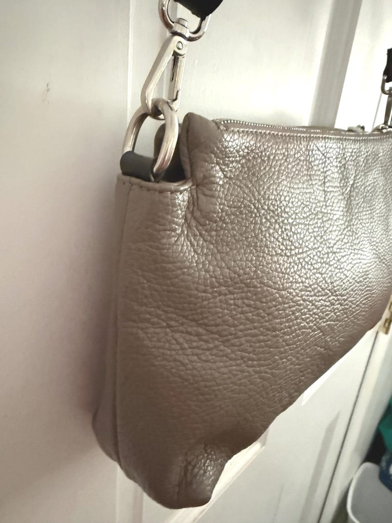 May include: A metallic taupe crossbody bag with a textured leather exterior. It has a silver-tone metal chain strap and hardware. The zipper is visible along the top edge. The bag is hanging on a white door.