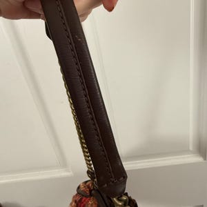 K&ouml;nnte beinhalten: Ein brauner Lederhandtaschengriff mit goldenen Kettenakzenten. Die Tasche hat einen gewebten Stoffk&ouml;rper in Rot-, Orange- und Cremet&ouml;nen. Der Griff wird von einer Person gehalten.