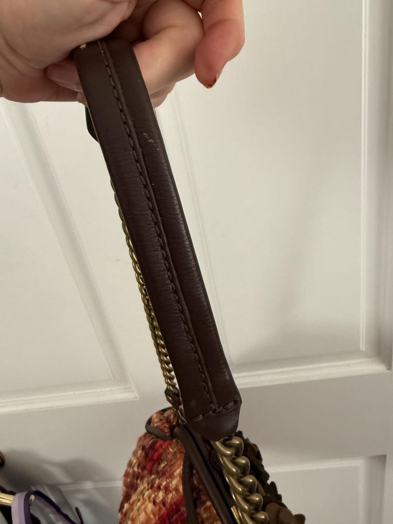 K&ouml;nnte beinhalten: Ein brauner Lederhandtaschengriff mit goldenen Kettenakzenten. Die Tasche hat einen gewebten Stoffk&ouml;rper in Rot-, Orange- und Cremet&ouml;nen. Der Griff wird von einer Person gehalten.