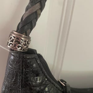 Puede incluir: Bolso de mano negro con asa trenzada y detalles met&aacute;licos plateados. El bolso tiene un patr&oacute;n texturizado similar a la piel de cocodrilo. La cremallera es visible a lo largo del borde superior.