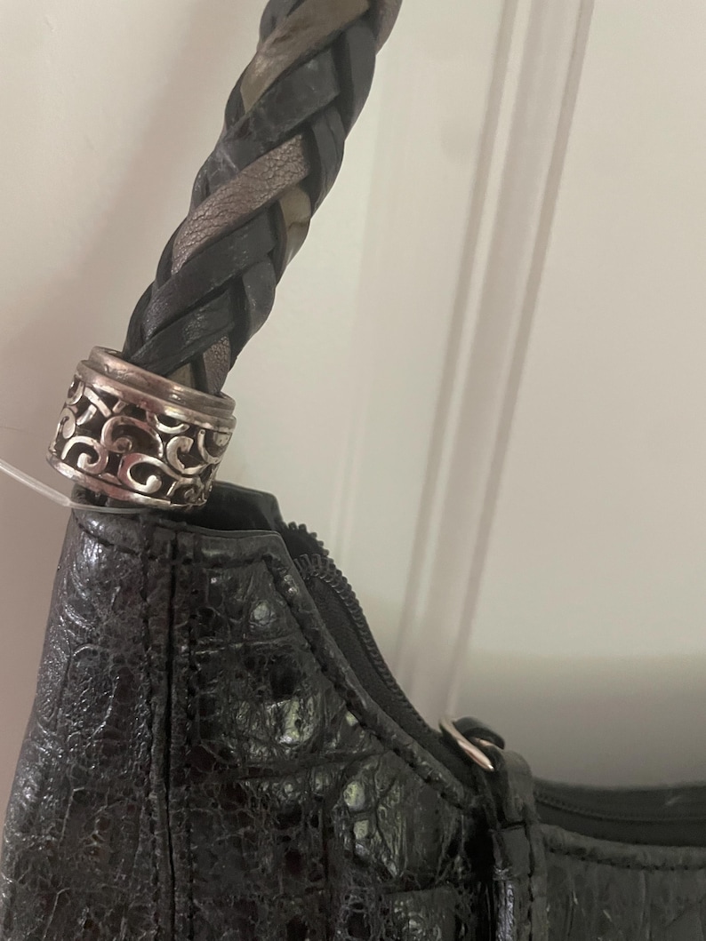 Puede incluir: Bolso de mano negro con asa trenzada y detalles met&aacute;licos plateados. El bolso tiene un patr&oacute;n texturizado similar a la piel de cocodrilo. La cremallera es visible a lo largo del borde superior.