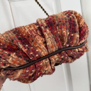 K&ouml;nnte beinhalten: Eine strukturierte Clutch in Rot-, Orange- und Braunt&ouml;nen mit dunkelbraunem Lederbesatz. Die Tasche hat ein gerafftes Design und einen Kettenriemen. Die Gesamt&auml;sthetik ist warm und herbstlich.