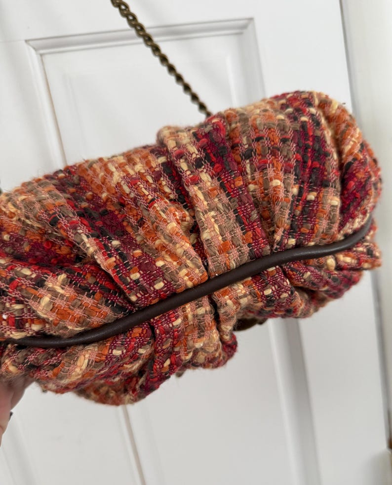 K&ouml;nnte beinhalten: Eine strukturierte Clutch in Rot-, Orange- und Braunt&ouml;nen mit dunkelbraunem Lederbesatz. Die Tasche hat ein gerafftes Design und einen Kettenriemen. Die Gesamt&auml;sthetik ist warm und herbstlich.