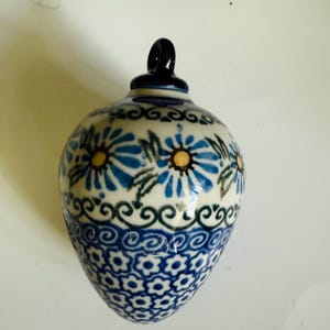 May include: A ceramic ornament with a blue and white floral design. The ornament has a teardrop shape with a black loop at the top. The design features blue flowers with yellow centers and green leaves. The bottom portion has a repeating blue floral pattern.
