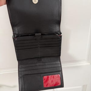 May include: Open black leather wallet with multiple card slots, a zippered compartment, and a clear ID window with a red interior. The wallet has a magnetic snap closure and a small metal accent.
