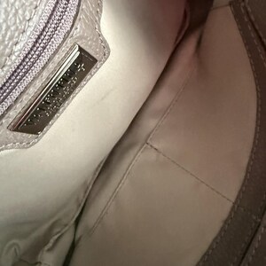 May include: Interior view of a light grey handbag. The inside is a light beige fabric with a stitched pocket. A silver metal tag with embossed text is visible near the zipper.