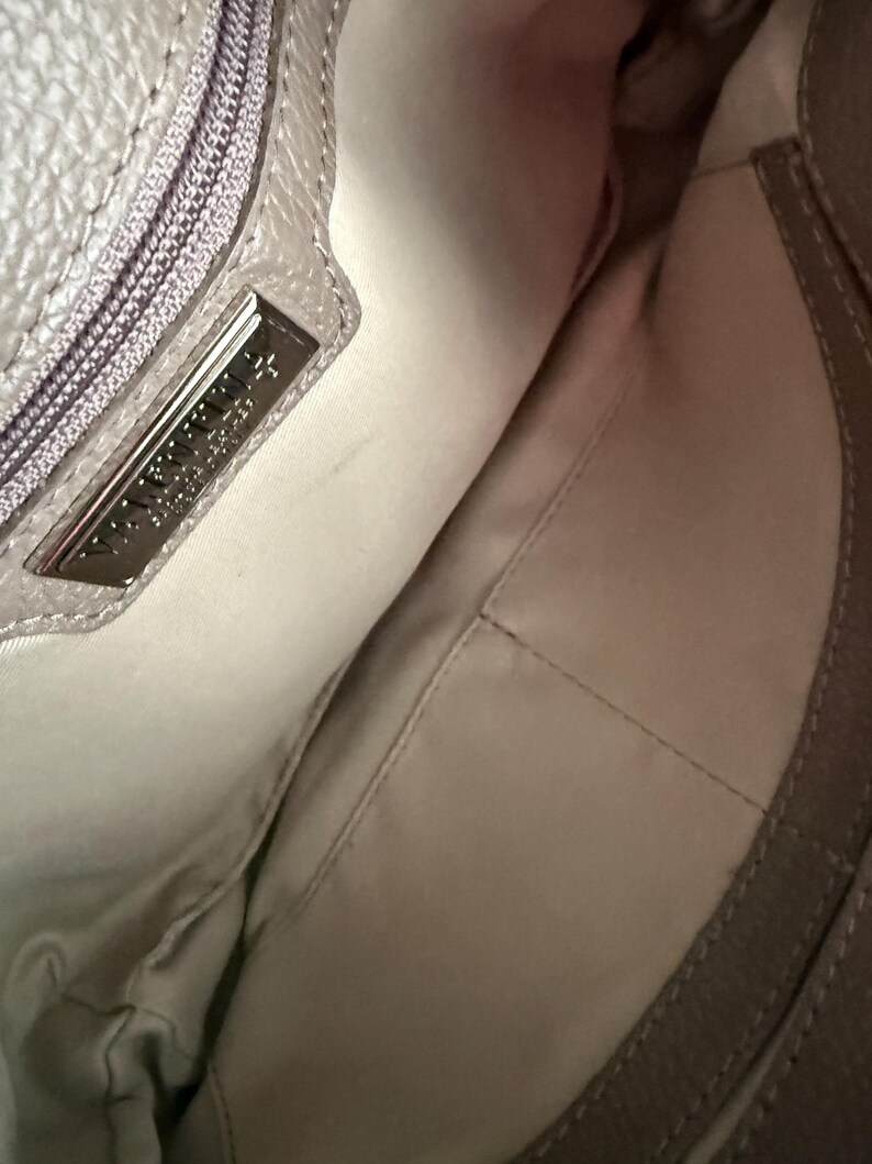 May include: Interior view of a light grey handbag. The inside is a light beige fabric with a stitched pocket. A silver metal tag with embossed text is visible near the zipper.