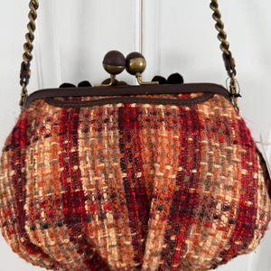 K&ouml;nnte beinhalten: Eine kleine, runde Handtasche mit einem gewebten Tweed-&Auml;u&szlig;eren in Rot-, Orange- und Beiget&ouml;nen. Die Tasche hat einen dunkelbraunen Rahmen, einen Schnallenverschluss mit zwei braunen und goldenen Kugeln und einen Kettenriemen.