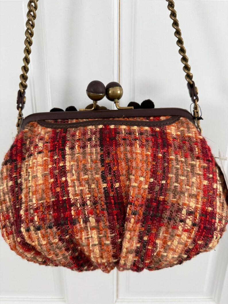 K&ouml;nnte beinhalten: Eine kleine, runde Handtasche mit einem gewebten Tweed-&Auml;u&szlig;eren in Rot-, Orange- und Beiget&ouml;nen. Die Tasche hat einen dunkelbraunen Rahmen, einen Schnallenverschluss mit zwei braunen und goldenen Kugeln und einen Kettenriemen.