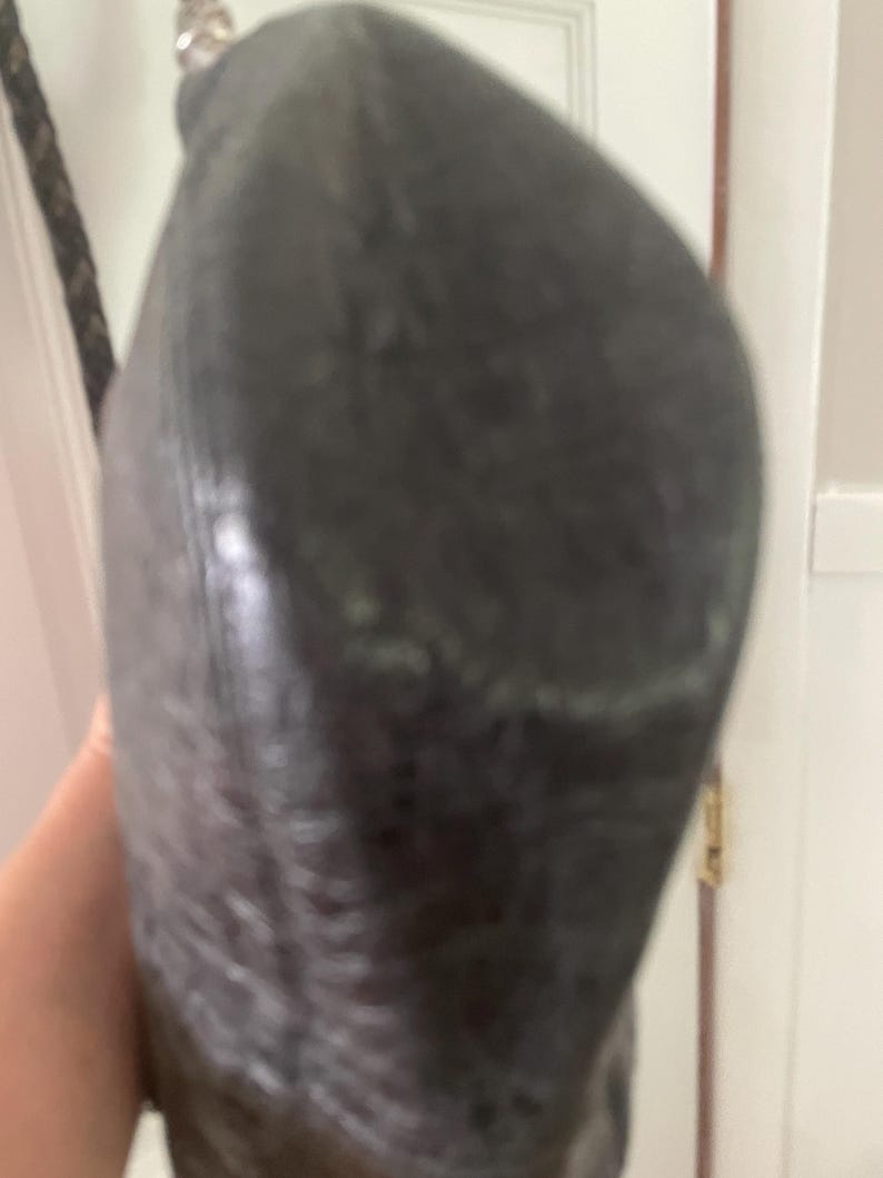 Puede incluir: Un bolso de mano gris oscuro texturizado con una correa trenzada. El bolso parece estar hecho de un material suave y tiene una forma redondeada. La correa es de color marr&oacute;n oscuro.