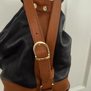May include: Black and brown leather backpack with adjustable straps and gold-tone hardware. The backpack features a drawstring closure and a brown leather base. The straps are brown leather with gold-tone buckles and decorative studs.