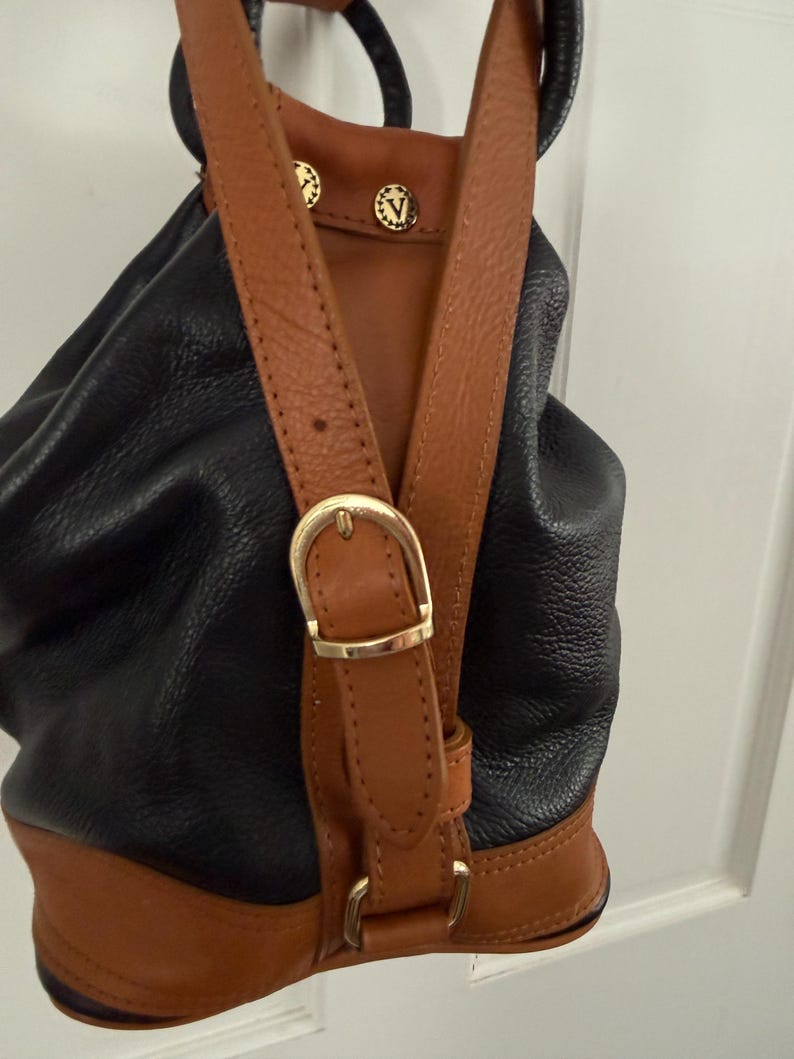 May include: Black and brown leather backpack with adjustable straps and gold-tone hardware. The backpack features a drawstring closure and a brown leather base. The straps are brown leather with gold-tone buckles and decorative studs.