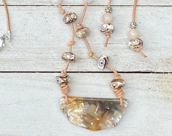 May include: A brown leather necklace with a large, irregular, agate pendant. The necklace features small, white and brown beads and silver accents. The necklace is displayed on a white wooden surface.