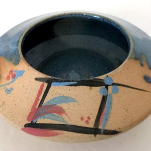 May include: A ceramic bowl with a blue glaze and abstract painted design in shades of red, blue, and black. The bowl has a wide, shallow shape and a textured surface.