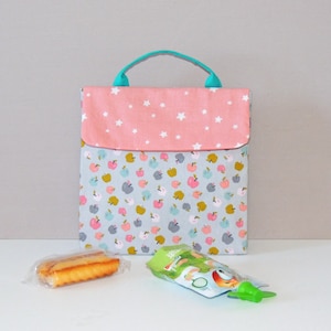 May include: A fabric lunch bag with a light blue background and a pattern of colorful apples. The flap is pink with white stars. It has a teal handle. A snack and a fruit pouch are in front of the bag.