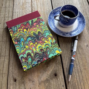May include: A rectangular notebook with a maroon fabric spine and a cover featuring a vibrant, multi-colored marbled pattern of yellow, green, blue, and red swirls. Beside it sits a blue and white ceramic teacup with black coffee and a ballpoint pen.
