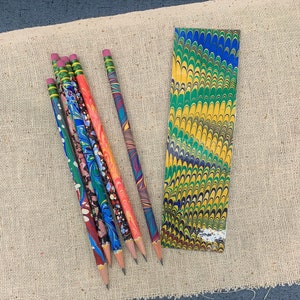 May include: A set of seven colorful pencils with marbled patterns. The pencils are arranged in a fan shape on a beige fabric background.