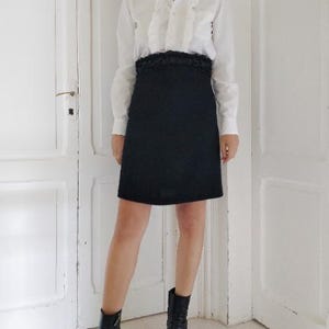 Può includere: Una camicia bianca con dettagli a balze e una gonna nera. La gonna ha un aspetto strutturato. L'outfit è completato da stivaletti neri con tacco a blocco. L'ambientazione è una stanza con porte bianche.