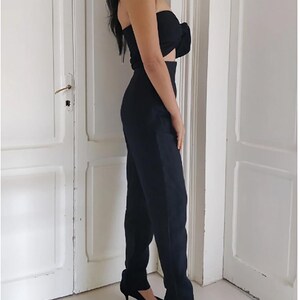 Puede incluir: Conjunto negro de dos piezas que consta de un top sin tirantes con detalle de lazo y pantalones de cintura alta con pernera recta y abertura en el tobillo. El conjunto se completa con tacones altos negros.