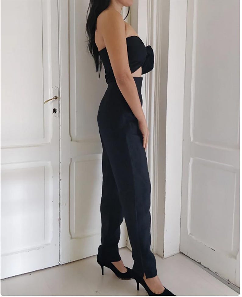 Puede incluir: Conjunto negro de dos piezas que consta de un top sin tirantes con detalle de lazo y pantalones de cintura alta con pernera recta y abertura en el tobillo. El conjunto se completa con tacones altos negros.