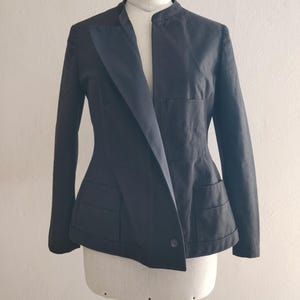 Può includere: Un blazer nero con un design asimmetrico unico. Il blazer presenta un colletto a revers, maniche lunghe e due tasche anteriori. È esposto su un manichino su uno sfondo bianco. Il blazer ha una chiusura con un solo bottone.