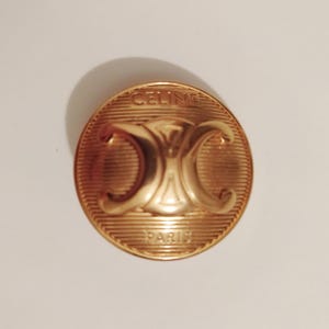 May include: A gold-coloured, circular button with the brand name "CELINE" arched across the top and "PARIS" at the bottom. The button features a central design of two interlocking "C" shapes.