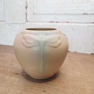 Könnte beinhalten: Eine matte, beige Keramikvase mit rundem Körper und weiter Öffnung. Die Außenseite zeigt ein erhabenes, geformtes Libellenrelief, das sich vertikal vom Hals bis zur Mitte des Gefäßes erstreckt und glatte Kurven aufweist.