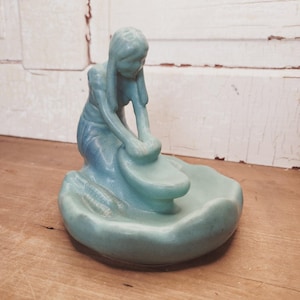 Könnte beinhalten: Eine hellblaue Keramikfigur, die eine kniende Person darstellt, die mit einem Steinwerkzeug auf einer runden Basis Getreide mahlt. Die Figur hat langes Haar und beugt sich über ein geschwungenes Becken, mit einer glatten, glänzenden Oberfläche.