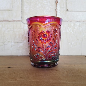 Puede incluir: Un vaso de vidrio iridiscente con un diseño floral. El vidrio presenta un girasol en el centro, rodeado de espigas de trigo en relieve y detalles ornamentados. Los colores cambian de rojo a naranja y verde.