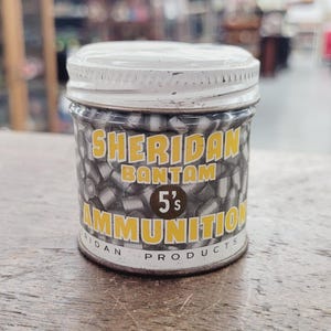May include: A small, cylindrical metal container with a white lid and a label that reads "Sheridan Bantam 5's AMMUNITION." The label is yellow and black. The container is filled with small, silver pellets.