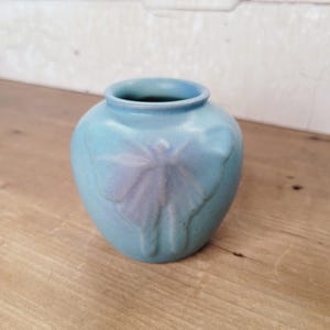 May include: A small, round, light blue ceramic vase with a matte finish. The exterior features a raised, embossed butterfly design centered on the front. The vase has a wide, circular opening at the top and a smooth, rounded body shape.